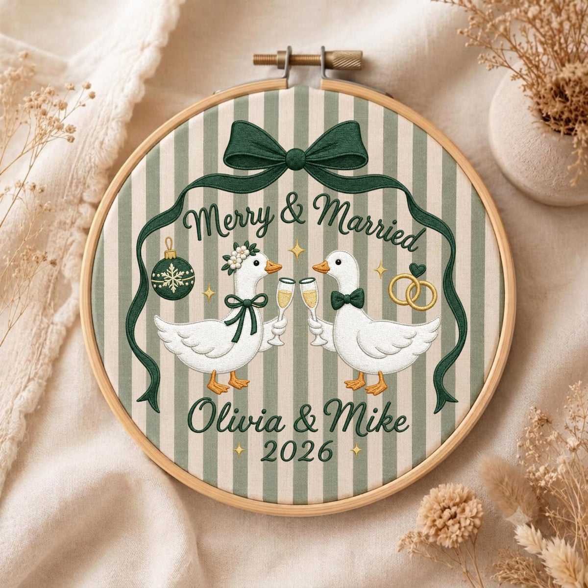 May include: A round wooden embroidery hoop featuring a striped fabric background with two white geese holding champagne flutes. The text reads Merry &amp; Married, Olivia &amp; Mike, and 2026. A dark green ribbon bow sits at the top with a green ornament.