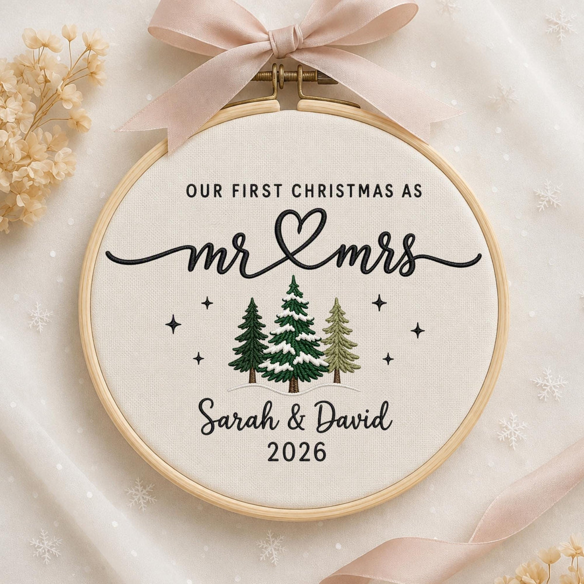 May include: A round wooden embroidery hoop containing cream-colored fabric with black embroidered text reading OUR FIRST CHRISTMAS AS mr &amp; mrs, Sarah &amp; David, and 2026. Three green pine trees and small black stars are stitched below.