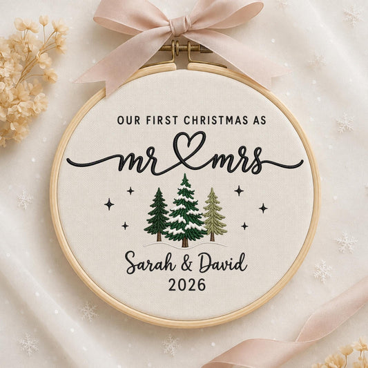 May include: A round wooden embroidery hoop containing cream-colored fabric with black embroidered text reading OUR FIRST CHRISTMAS AS mr &amp; mrs, Sarah &amp; David, and 2026. Three green pine trees and small black stars are stitched below.