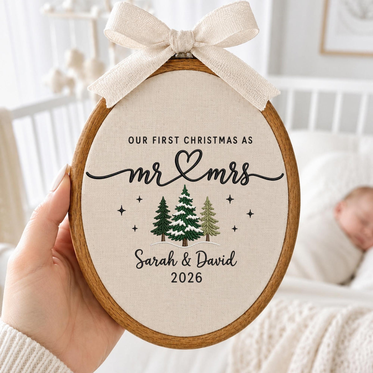May include: An oval wooden embroidery hoop containing cream-colored fabric with black embroidered text reading "OUR FIRST CHRISTMAS AS mr &amp; mrs" above a heart, three pine trees, the names "Sarah &amp; David", and the year "2026" with a beige bow.