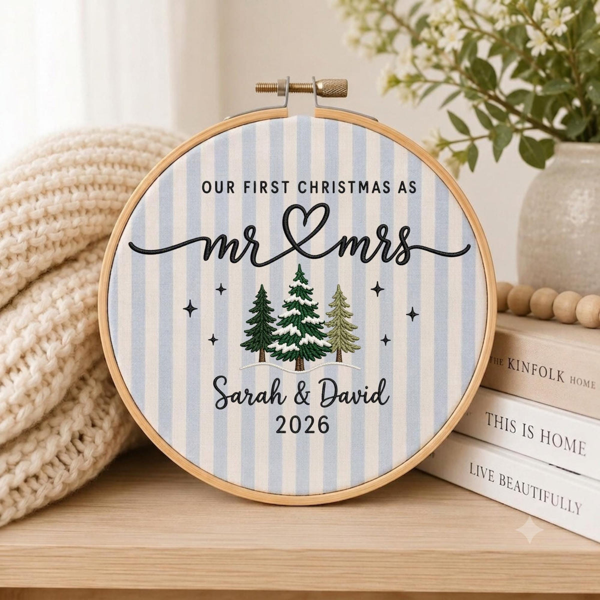 May include: A round wooden embroidery hoop containing fabric with light blue and white vertical stripes. The embroidery features the text "OUR FIRST CHRISTMAS AS mr &amp; mrs", "Sarah &amp; David", and "2026" in black, alongside three pine trees.