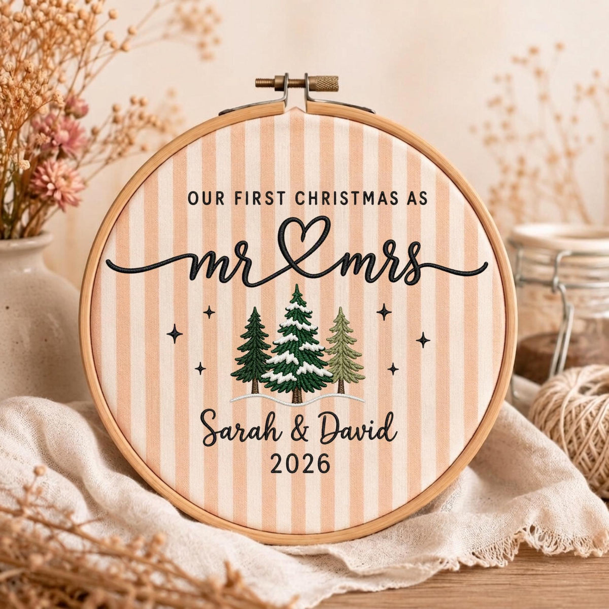May include: A round wooden embroidery hoop containing fabric with vertical beige stripes. The text reads "OUR FIRST CHRISTMAS AS mr &amp; mrs" with "Sarah &amp; David" and "2026" below. Three embroidered pine trees and small stars are centered.
