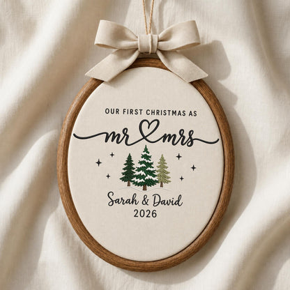 May include: An oval wooden embroidery hoop ornament featuring cream fabric with black embroidered text reading Our First Christmas as Mr &amp; Mrs, Sarah &amp; David, and 2026. Three embroidered pine trees and small stars sit below a beige ribbon bow.