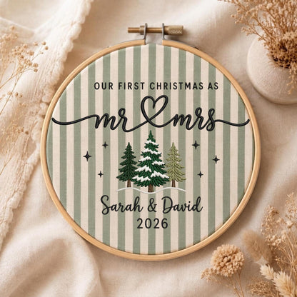 May include: A circular wooden embroidery hoop framing fabric with vertical sage green and white stripes. The embroidery features the black text "OUR FIRST CHRISTMAS AS mr &amp; mrs", "Sarah &amp; David", and "2026", with three stitched pine trees.