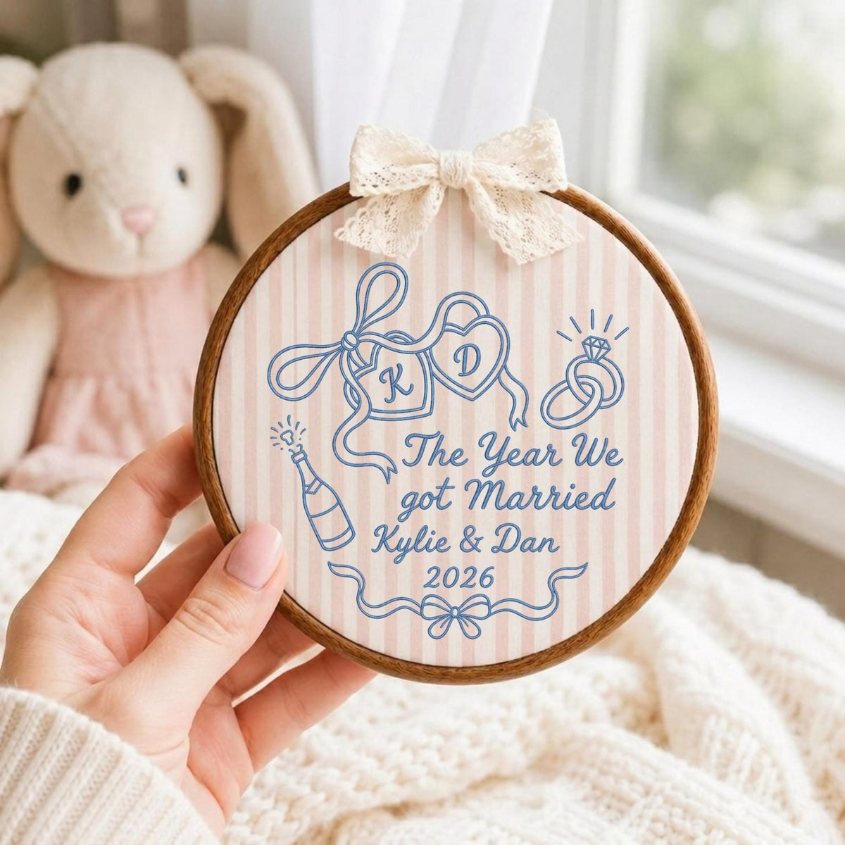 May include: A round wooden embroidery hoop containing fabric with a light pink and white vertical stripe pattern. The fabric features blue embroidered designs including two hearts with letters K and D, a champagne bottle, and rings. Text reads: The Year We got Married Kylie &amp; Dan 2026. A cream lace bow is attached to the top.