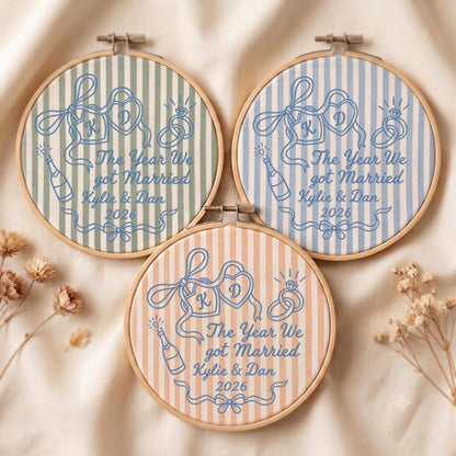 May include: Three circular wooden embroidery hoops featuring striped fabric in sage green, light blue, and peach. Each hoop displays blue embroidered designs of two hearts with initials K and D, wedding rings, a champagne bottle, and the text: The Year We got Married, Kylie &amp; Dan, 2026.