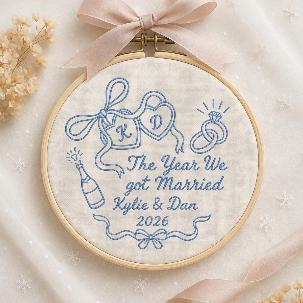 May include: A circular wooden embroidery hoop containing off-white fabric with blue stitched designs including two hearts with letters K and D, two interlocking rings, a champagne bottle, and the text The Year We got Married Kylie &amp; Dan 2026.