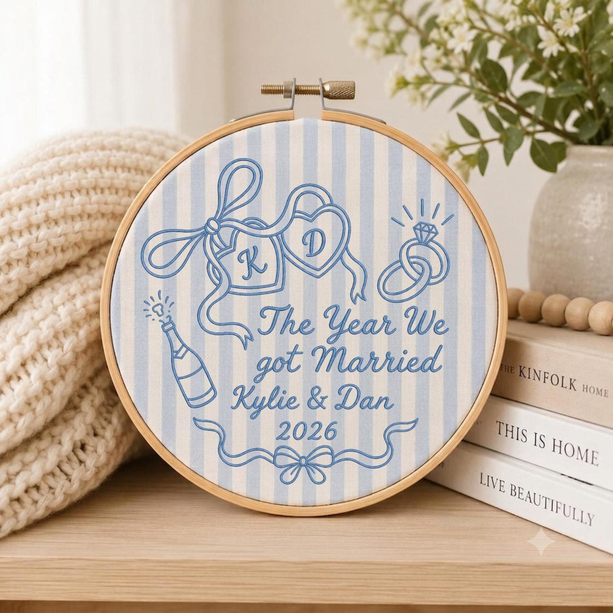 May include: A round wooden embroidery hoop containing light blue and white striped fabric with blue embroidered designs including two hearts with initials K and D, a champagne bottle, a diamond ring, and the text The Year We got Married Kylie &amp; Dan 2026.