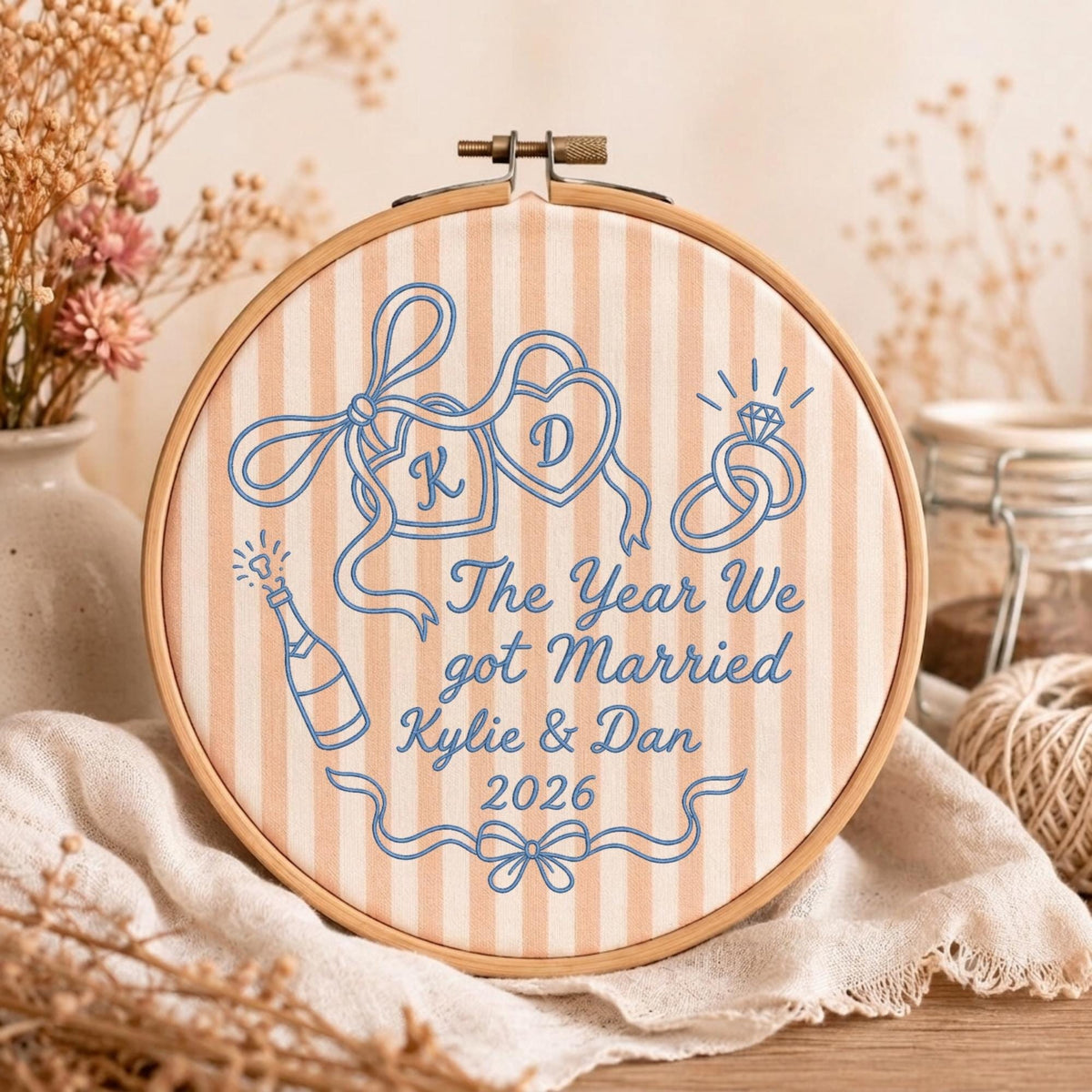 May include: A round wooden embroidery hoop containing fabric with vertical beige and white stripes. Blue embroidery features two heart-shaped locks with initials K and D, two wedding rings, a champagne bottle, and the text The Year We got Married Kylie &amp; Dan 2026.