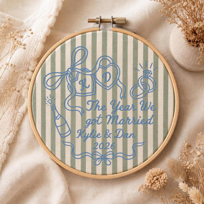 May include: A circular wooden embroidery hoop containing sage green and white striped fabric with blue thread embroidery. The design features two heart-shaped locks with initials K and D, a champagne bottle, and rings, with the text The Year We got Married Kylie &amp; Dan 2026.