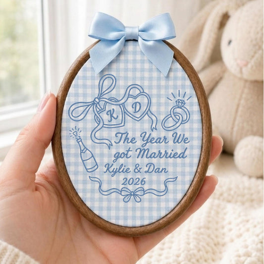 May include: An oval wooden embroidery hoop frame containing light blue and white gingham fabric. The fabric features blue embroidered designs of two hearts with letters K and D, a ring, a champagne bottle, and text: The Year We got Married Kylie &amp; Dan 2026. A light blue satin ribbon bow is attached to the top.
