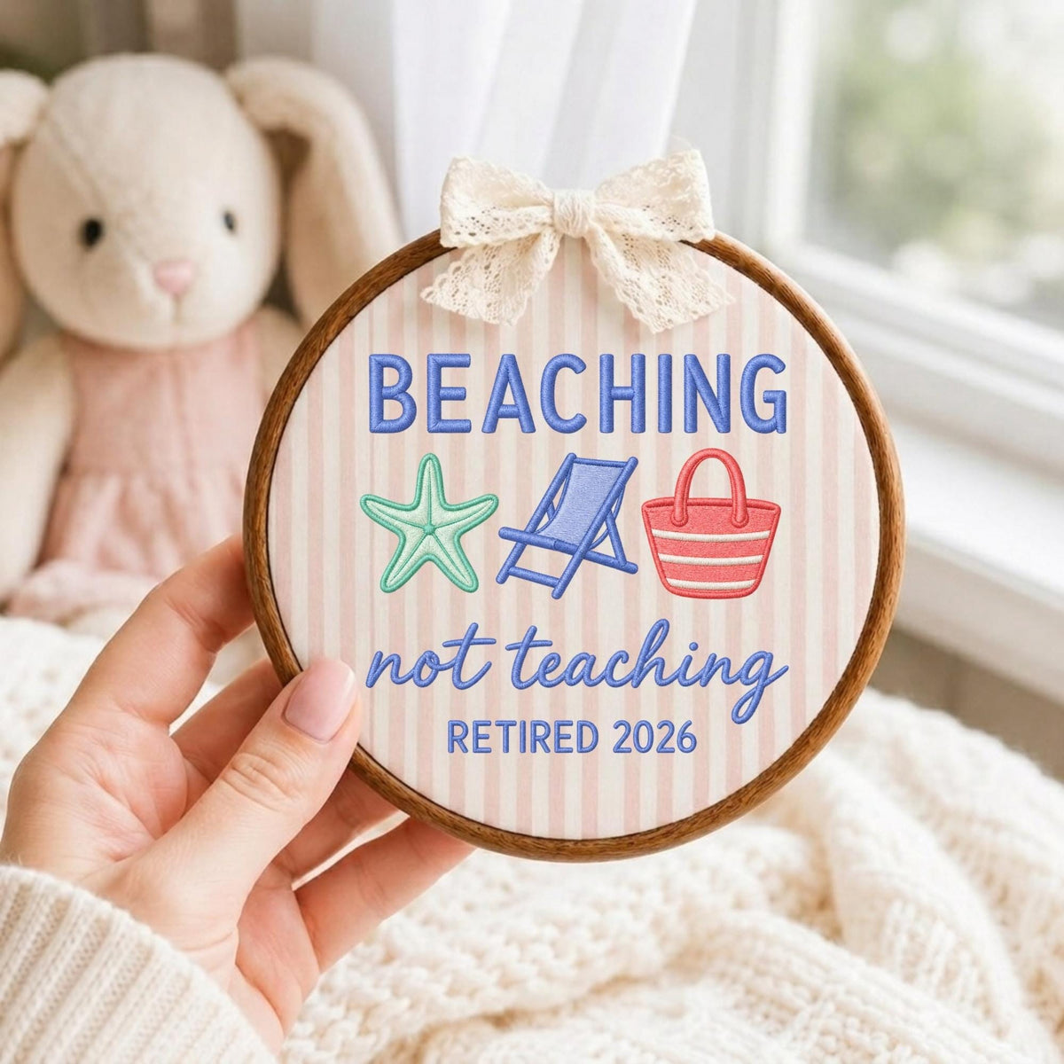 May include: A circular wooden embroidery hoop containing fabric with vertical light pink stripes. The embroidery features the blue text BEACHING not teaching and RETIRED 2026, with a green starfish, a blue beach chair, and a red beach bag.