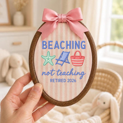 May include: An oval embroidery hoop containing fabric with vertical pink and white stripes. The text reads BEACHING not teaching RETIRED 2026 in blue. Embroidered icons include a mint starfish, a blue beach chair, and a pink beach bag. A pink satin bow is attached to the top.