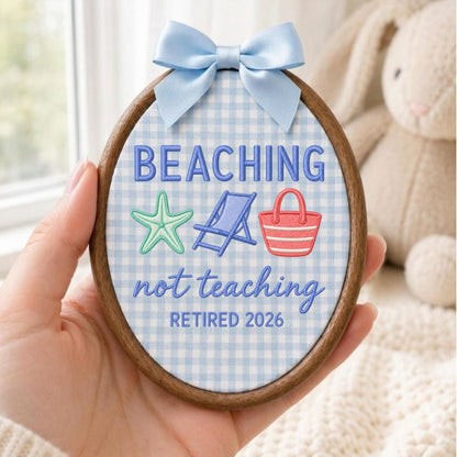 May include: An oval embroidery hoop featuring a light blue and white gingham fabric background. The text reads BEACHING not teaching RETIRED 2026 in blue thread. Embroidered icons include a mint green starfish, a blue beach chair, and a red tote bag.