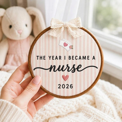May include: A circular wooden embroidery hoop frame containing fabric with vertical light pink stripes. The text reads THE YEAR I BECAME A nurse 2026, featuring a nurse cap icon with a pink heart and a small flower, plus a cream lace bow.