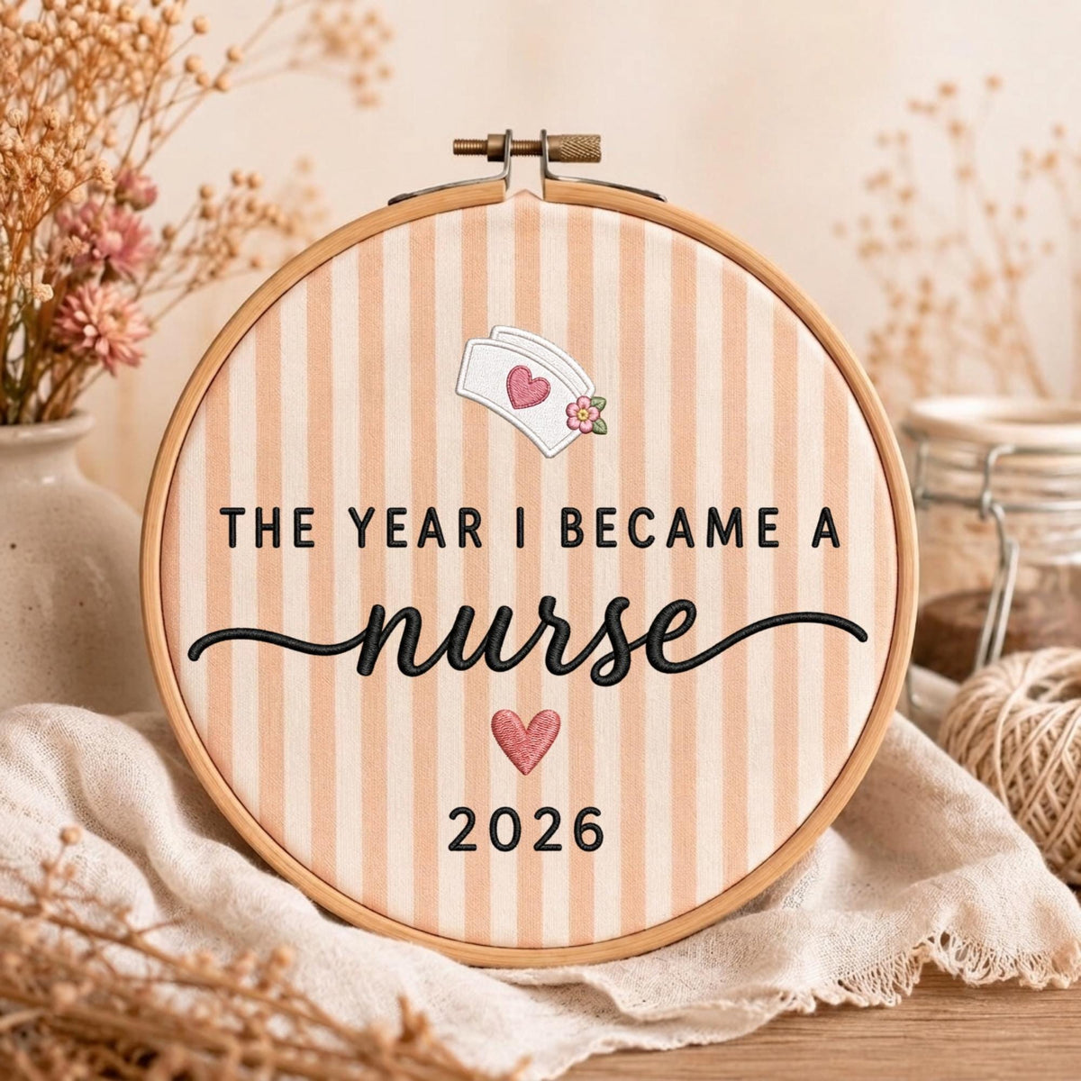 May include: A circular wooden embroidery hoop containing fabric with vertical peach and white stripes. The text reads THE YEAR I BECAME A nurse 2026. It features an embroidered white nurse cap with a pink heart and a small pink flower, plus a pink heart.