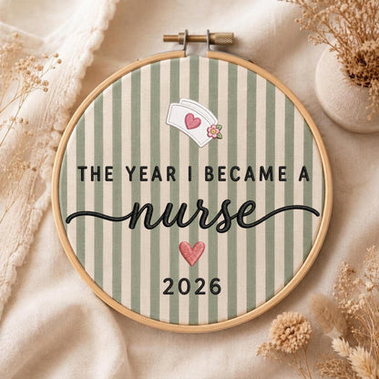 May include: A circular wooden embroidery hoop containing fabric with vertical sage green and cream stripes. The text reads THE YEAR I BECAME A nurse 2026. It features an embroidered white nurse cap with a pink heart, and a pink heart below the text.