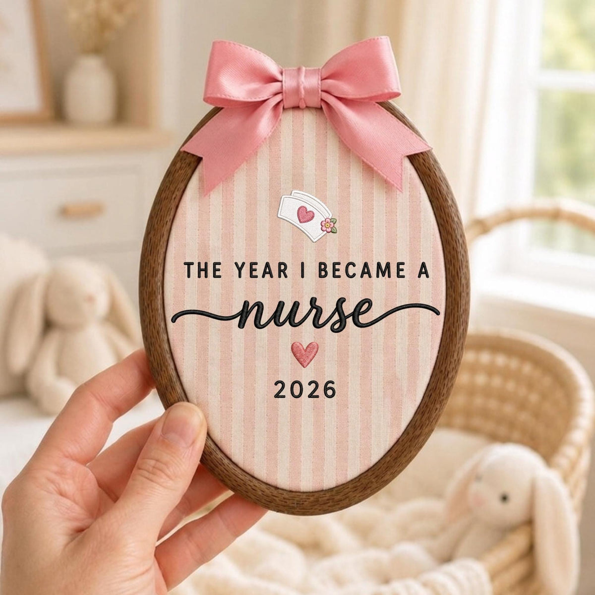 May include: An oval wooden embroidery hoop containing fabric with vertical pink and white stripes. A pink satin bow is attached to the top. The text reads THE YEAR I BECAME A nurse 2026, with a nurse cap icon and two small pink hearts.