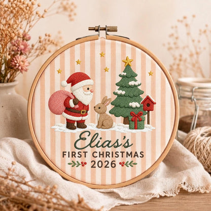 May include: A circular wooden embroidery hoop framing a fabric piece with vertical beige stripes. It features embroidered designs of Santa Claus with a pink sack, a brown rabbit, a green Christmas tree, and a red birdhouse. Text reads: Elia's FIRST CHRISTMAS 2026.
