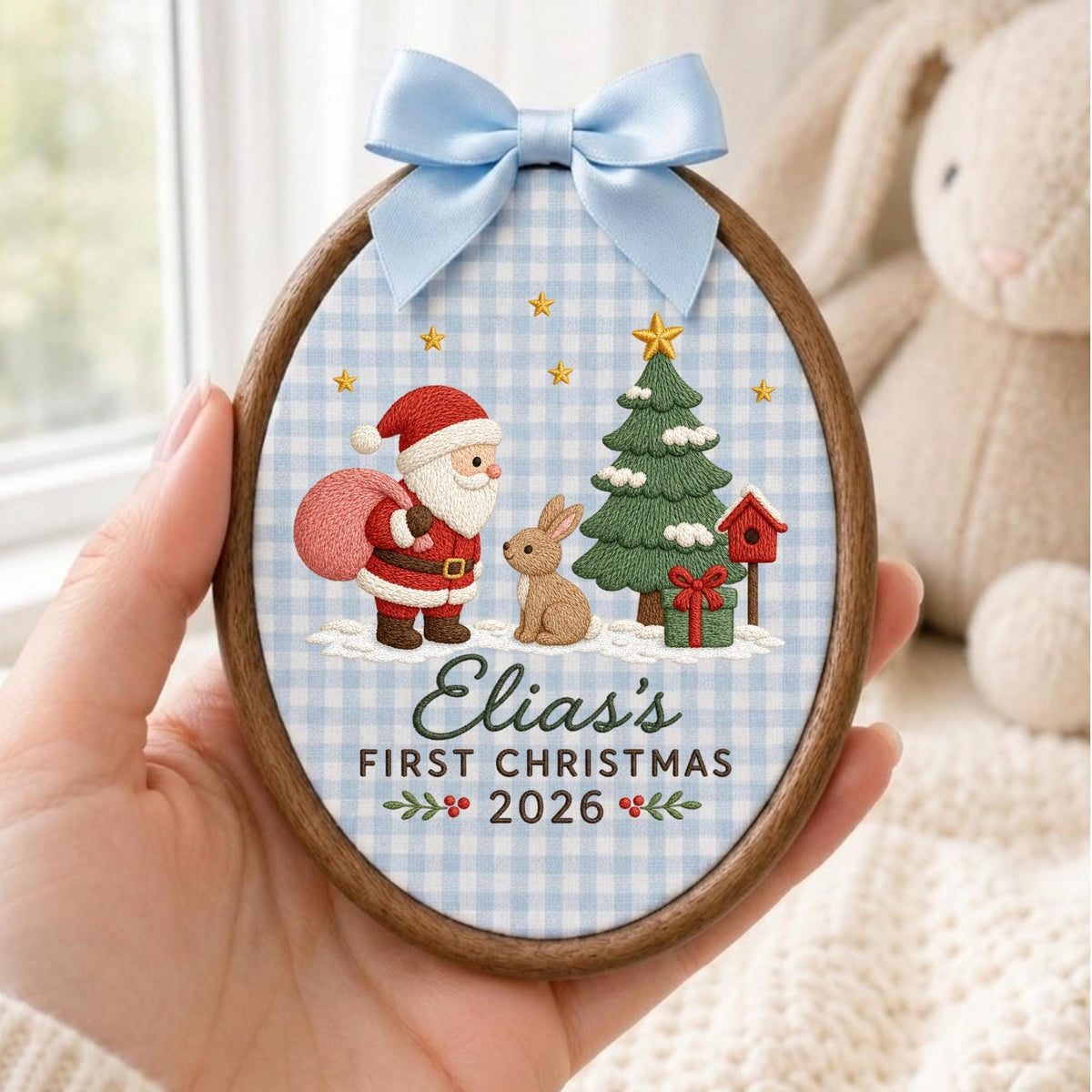 May include: An oval wooden embroidery hoop ornament featuring a light blue gingham fabric background. The embroidery depicts Santa Claus with a pink sack, a brown rabbit, a green Christmas tree, and a red birdhouse. Text reads: Elia's FIRST CHRISTMAS 2026. A light blue satin bow is attached to the top.