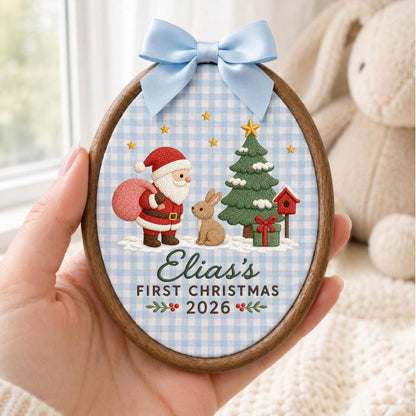 May include: An oval wooden embroidery hoop ornament featuring a light blue gingham fabric background. The embroidery depicts Santa Claus with a pink sack, a brown rabbit, a green Christmas tree, and a red birdhouse. Text reads: Elia's FIRST CHRISTMAS 2026. A light blue satin bow is attached to the top.