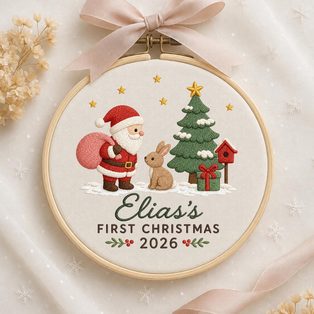 May include: A circular embroidery hoop featuring a festive scene with Santa Claus, a brown rabbit, a green Christmas tree, and a red birdhouse. The text reads Elia's FIRST CHRISTMAS 2026. A light pink ribbon is tied at the top of the wooden frame.