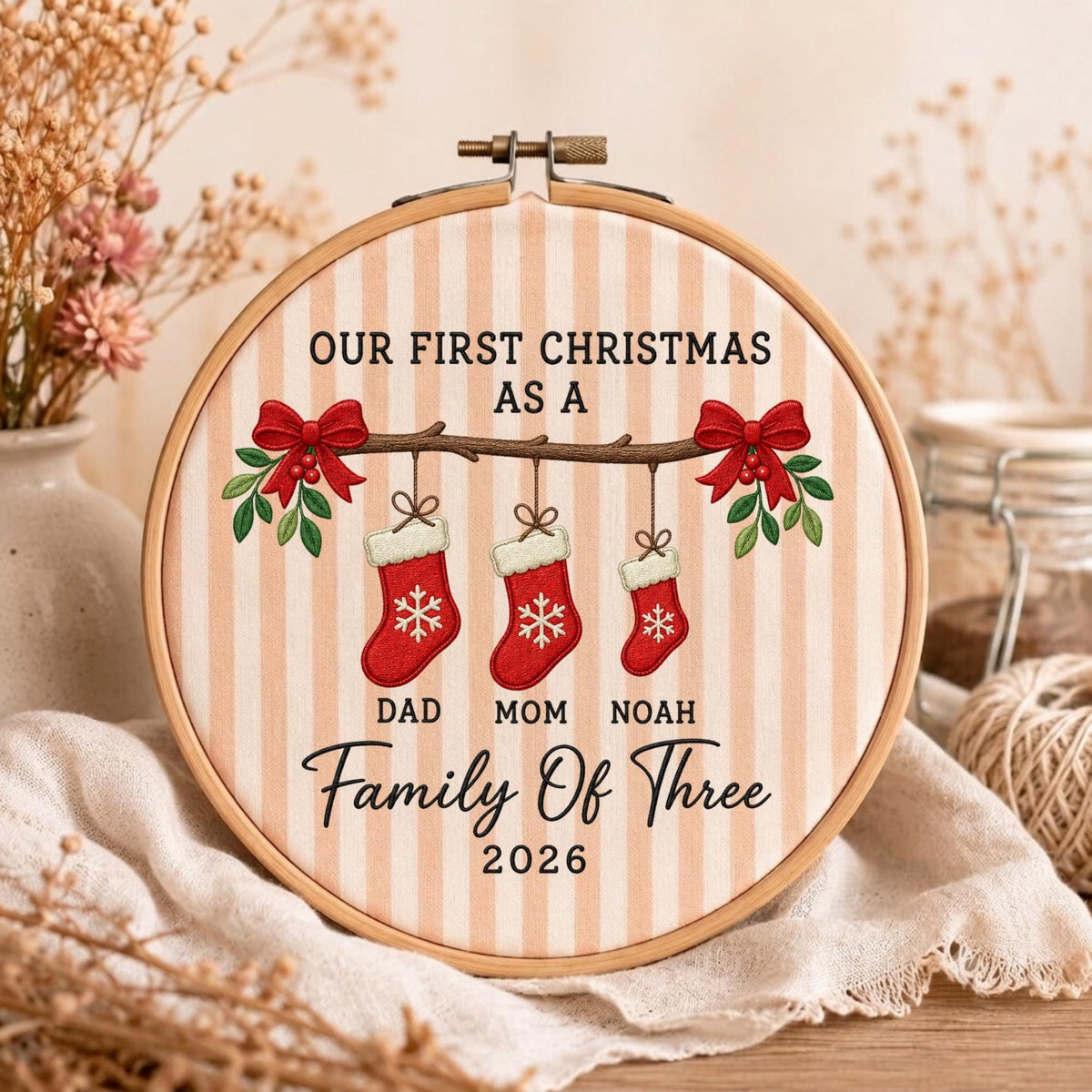 May include: A circular wooden embroidery hoop containing fabric with vertical beige stripes. The design features the text OUR FIRST CHRISTMAS AS A FAMILY OF THREE 2026, with three red stockings hanging from a branch, labeled DAD, MOM, and NOAH.