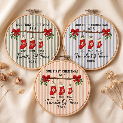 May include: Three circular embroidery hoops featuring striped fabric in sage green, light blue, and peach. Each displays a branch with red bows, three red stockings, and the text: OUR FIRST CHRISTMAS AS A FAMILY OF THREE 2026 DAD MOM NOAH.