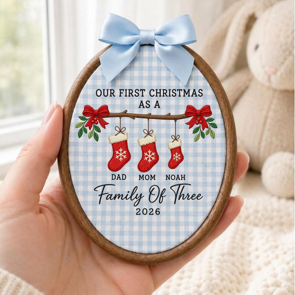 May include: An oval wooden embroidery hoop ornament featuring a light blue and white gingham fabric background. It displays the text "OUR FIRST CHRISTMAS AS A FAMILY OF THREE 2026" with three red stockings labeled "DAD", "MOM", and "NOAH" and a blue bow.