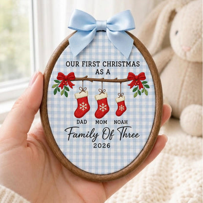 May include: An oval wooden embroidery hoop ornament featuring a light blue and white gingham fabric background. It displays the text "OUR FIRST CHRISTMAS AS A FAMILY OF THREE 2026" with three red stockings labeled "DAD", "MOM", and "NOAH" and a blue bow.
