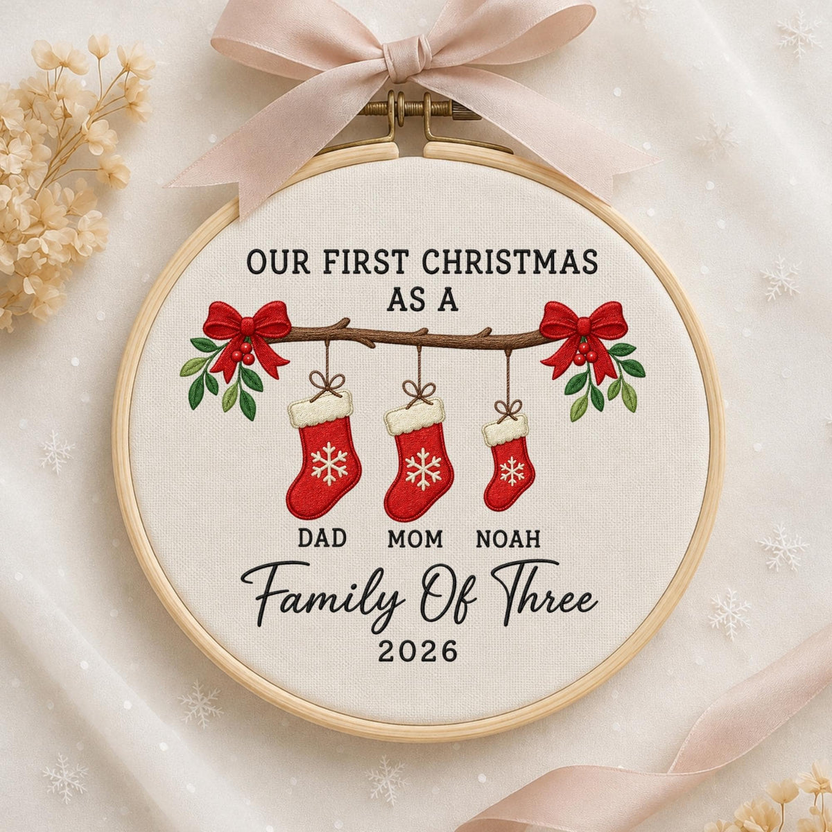 May include: A round wooden embroidery hoop containing white fabric with a design featuring three red Christmas stockings hanging from a branch. The text reads: OUR FIRST CHRISTMAS AS A DAD MOM NOAH Family Of Three 2026. A light pink ribbon is tied at the top.