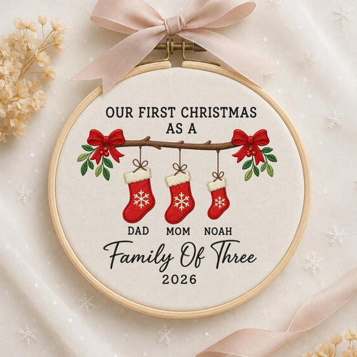 May include: A round wooden embroidery hoop containing white fabric with a design featuring three red Christmas stockings hanging from a branch. The text reads: OUR FIRST CHRISTMAS AS A DAD MOM NOAH Family Of Three 2026. A light pink ribbon is tied at the top.