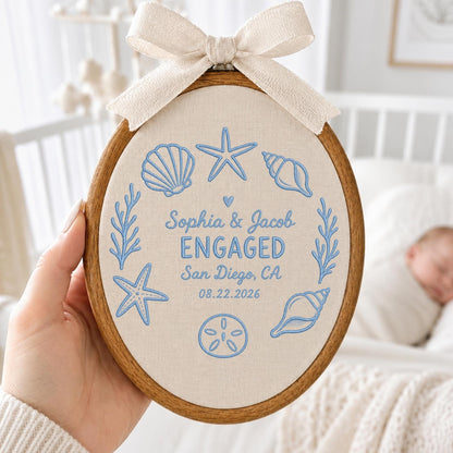 May include: An oval wooden embroidery hoop containing light-colored fabric with blue embroidered seashells, starfish, and coral. The text reads Sophia &amp; Jacob ENGAGED San Diego, CA 08.22.2026. A beige fabric bow is attached to the top of the hoop.