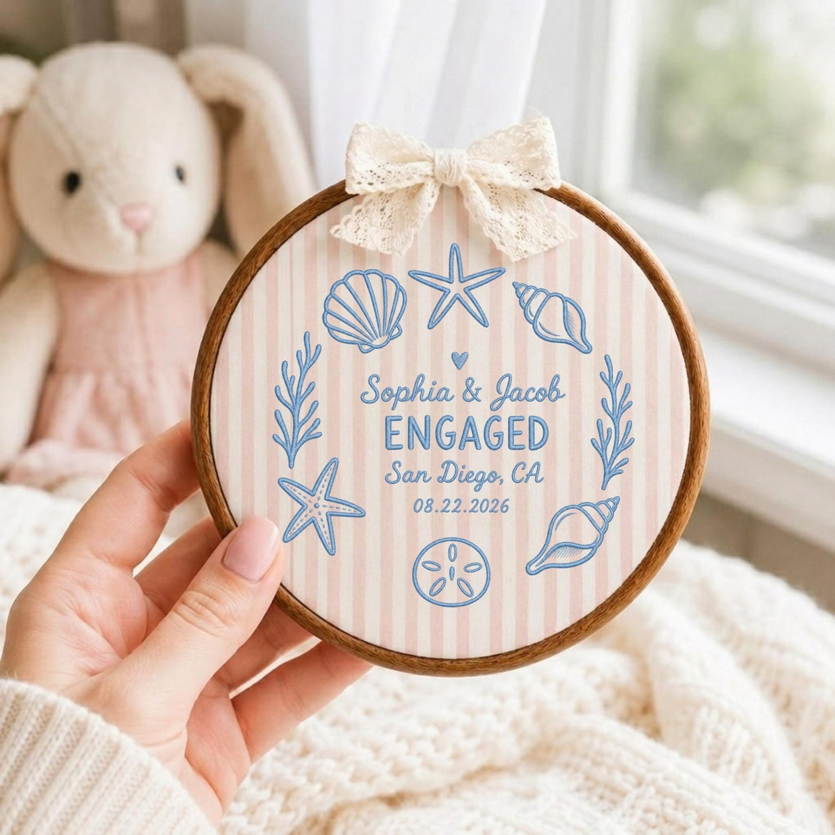 May include: A circular wooden embroidery hoop containing fabric with light pink vertical stripes. The fabric features blue embroidered seashells, starfish, and seaweed surrounding the text: Sophia &amp; Jacob ENGAGED San Diego, CA 08.22.2026.