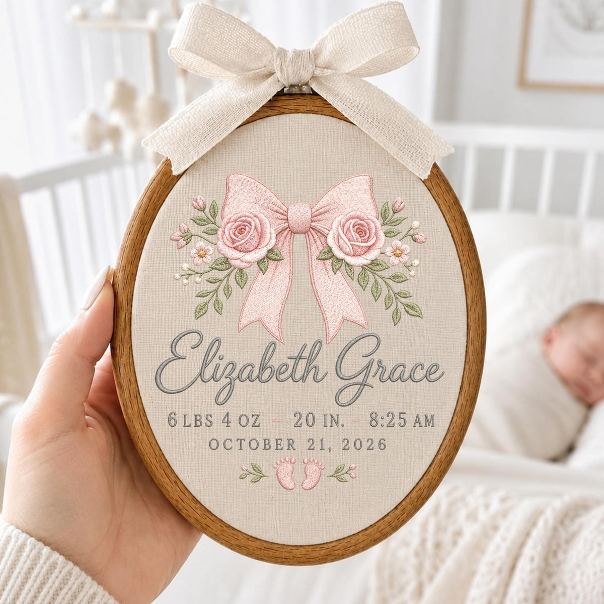 May include: An oval wooden embroidery hoop containing beige fabric with a pink embroidered bow and roses. The text reads Elizabeth Grace, 6 lbs 4 oz, 20 in, 8:25 am, October 21, 2026, with small baby footprints. A cream-colored fabric bow is tied at the top.