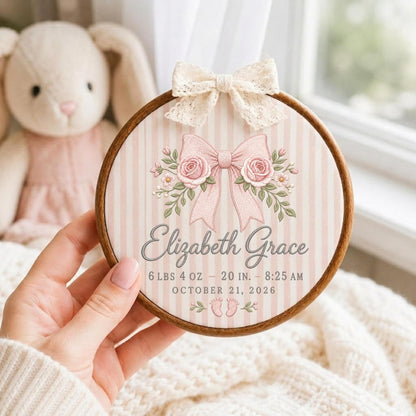 May include: A circular wooden embroidery hoop frame containing a fabric panel with a pink and white striped pattern. The design features a large pink bow with roses, the name Elizabeth Grace, birth details 6 lbs 4 oz, 20 in, 8:25 am, October 21, 2026, and small baby footprints, topped with a cream lace bow.