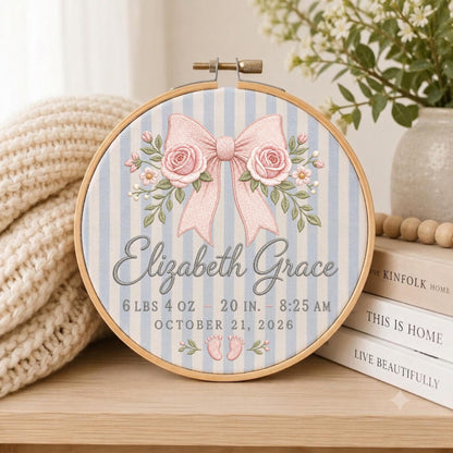 May include: A circular wooden embroidery hoop containing fabric with light blue and white vertical stripes. The design features an embroidered pink bow with roses, the name Elizabeth Grace, birth details 6 lbs 4 oz, 20 in, 8:25 am, October 21, 2026, and baby feet.