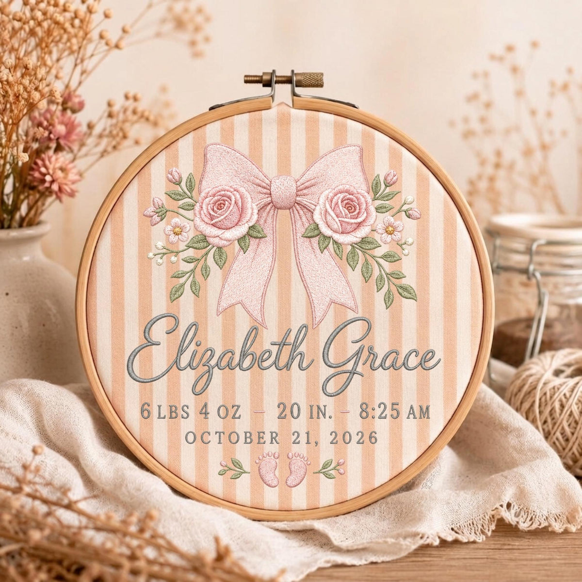 May include: A circular wooden embroidery hoop containing fabric with vertical beige stripes. The design features an embroidered pink bow with roses, the name Elizabeth Grace, birth details 6 lbs 4 oz, 20 in, 8:25 am, October 21, 2026, and baby footprints.