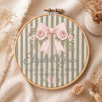 May include: A circular wooden embroidery hoop containing fabric with vertical sage green and white stripes. The embroidery features a large pink bow, two pink roses, the name Elizabeth Grace, and birth details: 6 lbs 4 oz, 20 in, 8:25 AM, October 21, 2026, and two small pink baby footprints.