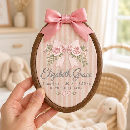 May include: An oval wooden embroidery hoop frame containing fabric with pink vertical stripes. The fabric features a pink satin bow at the top, a central illustration of a bow with roses, and the text Elizabeth Grace, 6 lbs 4 oz, 20 in, 8:25 AM, October 21, 2026, and two small footprints.