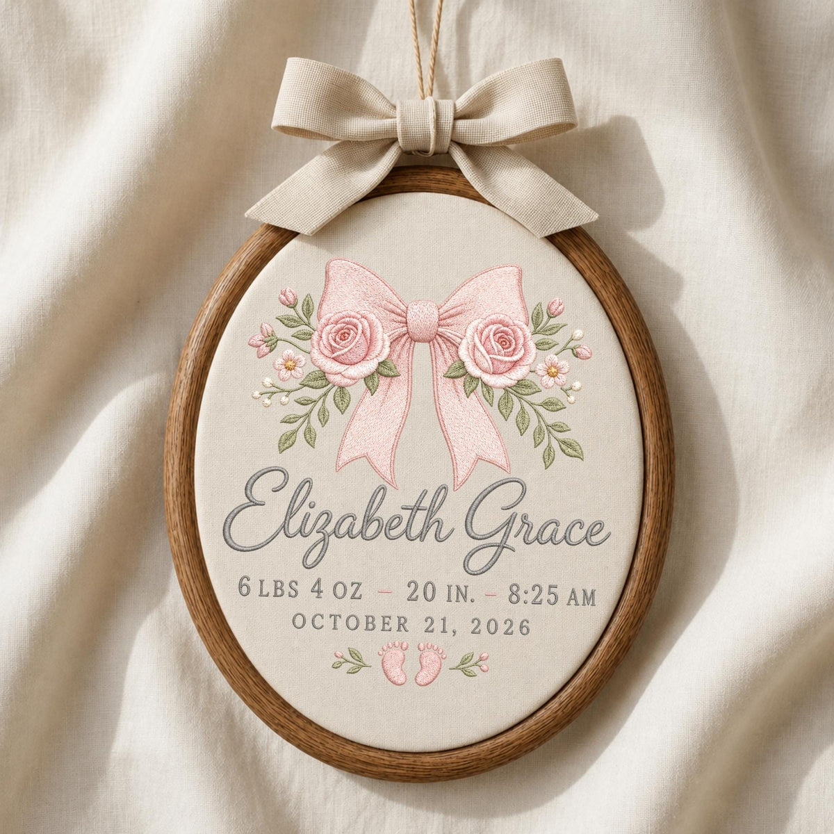 May include: An oval wooden embroidery hoop containing cream fabric with a beige ribbon bow at the top. The embroidery features a pink bow, two roses, and green leaves. Text reads: Elizabeth Grace, 6 lbs 4 oz - 20 in. - 8:25 AM, October 21, 2026, with two small pink baby footprints.