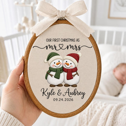 May include: An oval wooden embroidery hoop containing fabric with a printed design of two snowmen wearing green and red scarves and hats. The text reads "OUR FIRST CHRISTMAS AS mr &amp; mrs", "Kyle &amp; Aubrey", and "09.24.2026". A beige ribbon bow is attached to the top.