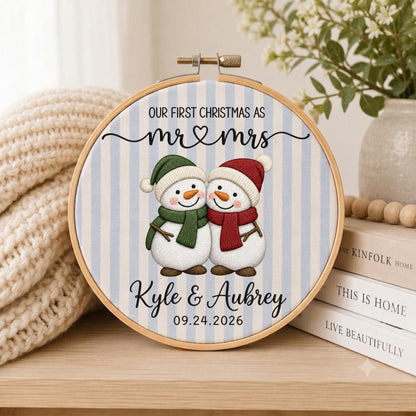 May include: A circular embroidery hoop containing fabric with light blue and white vertical stripes. The text reads OUR FIRST CHRISTMAS AS mr &amp; mrs Kyle &amp; Aubrey 09.24.2026. Two snowmen are centered, one wearing a green hat and scarf, one in red.