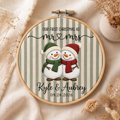 May include: A round wooden embroidery hoop framing fabric with vertical sage green stripes. The design features two snowmen with orange carrot noses, one wearing a green hat and scarf, the other a red hat and scarf. Text reads: OUR FIRST CHRISTMAS AS mr &amp; mrs Kyle &amp; Aubrey 09.24.2026.