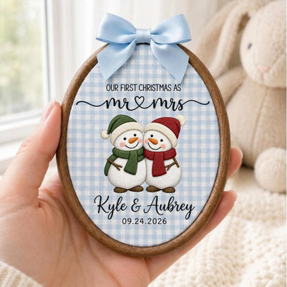 May include: An oval wooden embroidery hoop ornament featuring a light blue gingham fabric background. It displays two snowmen with scarves, the text "OUR FIRST CHRISTMAS AS mr &amp; mrs", the names "Kyle &amp; Aubrey", and the date "09.24.2026" with a light blue bow.