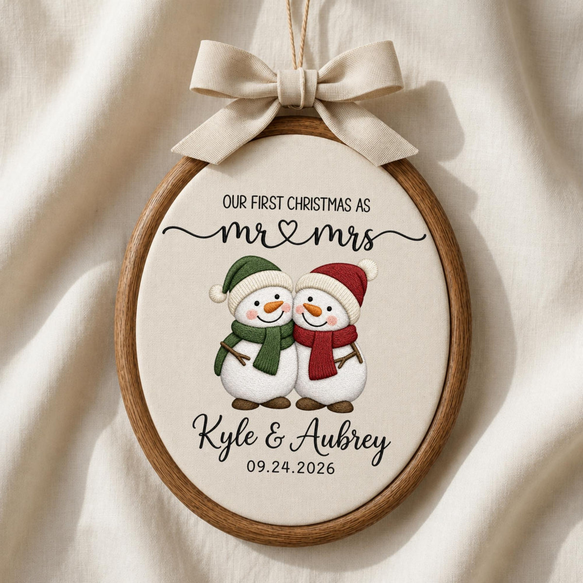May include: An oval wooden embroidery hoop ornament featuring a cream fabric center with two snowmen wearing green and red scarves and hats. The text reads: OUR FIRST CHRISTMAS AS mr &amp; mrs, Kyle &amp; Aubrey, 09.24.2026. A beige ribbon bow is attached.