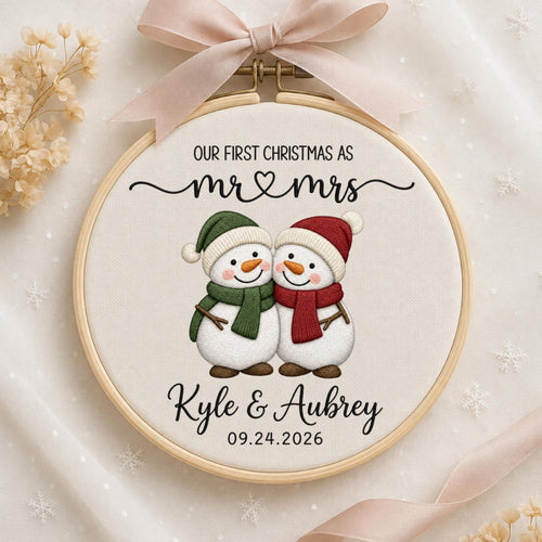 May include: A round wooden embroidery hoop containing white fabric with a printed design of two snowmen wearing green and red scarves and hats. The text reads: OUR FIRST CHRISTMAS AS mr &amp; mrs, Kyle &amp; Aubrey, and the date 09.24.2026.