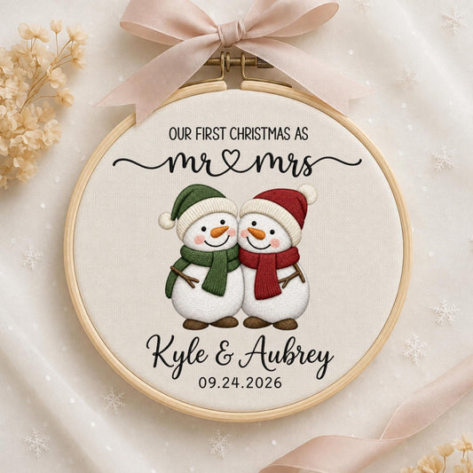 May include: A round wooden embroidery hoop containing white fabric with a printed design of two snowmen wearing green and red scarves and hats. The text reads: OUR FIRST CHRISTMAS AS mr &amp; mrs, Kyle &amp; Aubrey, and the date 09.24.2026.