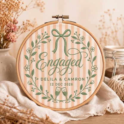 May include: A circular wooden embroidery hoop containing fabric with vertical peach and white stripes. The embroidery features a green floral wreath, a bow, two champagne flutes, and the text Engaged, Delila &amp; Camron, 12 Dec 2026.