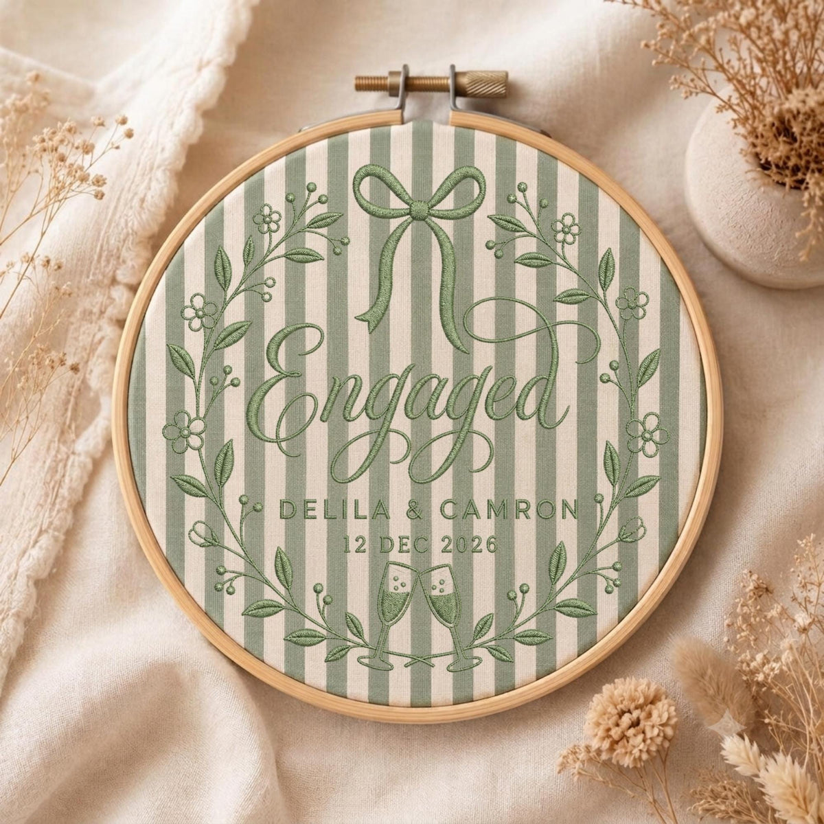 May include: A round wooden embroidery hoop containing fabric with sage green and white vertical stripes. The embroidery features a green floral wreath, a bow, two champagne flutes, and the text Engaged, Delila &amp; Camron, 12 Dec 2026.